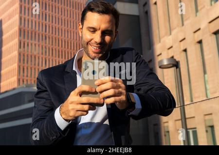 Bearded male entrepreneur in formal wear looking away while browsing on mobile phone standing on sunny street near building Stock Photo