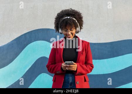 Happy African American Woman with Afro hairstyle and eyeglasses wearing black turtleneck and red jacket listening to podcast in wireless headphones wh Stock Photo