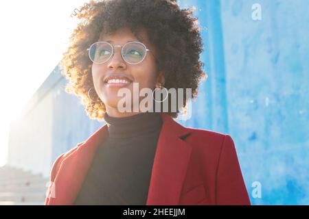 From below of smiling black female with Afro hairstyle standing near light blue stone fence and looking away Stock Photo