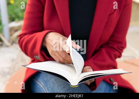 Crop anonymous female in red jacket and jeans sitting on bench and flipping through pages while reading book Stock Photo