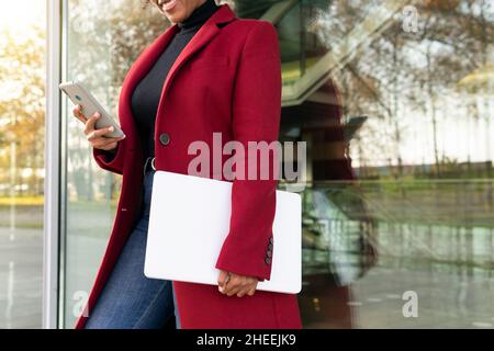 Side view of cropped unrecognizable African American Woman with laptop leaning on glass window showcase and messaging on smartphone Stock Photo