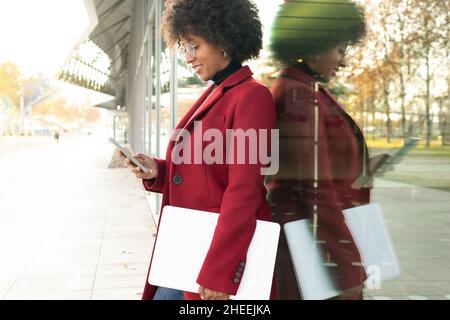 Side view of African American Woman with Afro hairstyle and laptop leaning on glass window showcase and messaging on smartphone Stock Photo