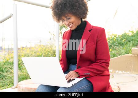 Positive young black Woman in eyeglasses wearing formal red jacket and jeans sitting on bench and checking email on laptop Stock Photo