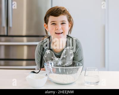 Cheerful kid in apron standing in light kitchen near table with bowl of flour and glass of water while preparing for cooking Stock Photo