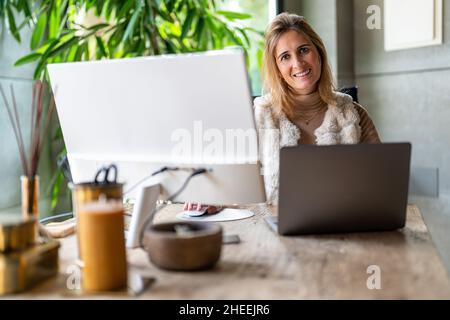 Cheerful female worker typing on laptop while looking at camera in light modern workspace with green plant during workday Stock Photo