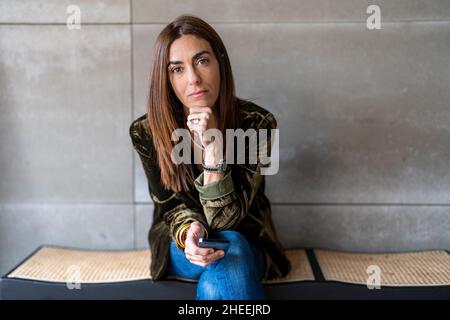 Focused female entrepreneur with cellphone while sitting on bench at gray wall in light modern office during workday Stock Photo