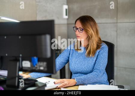 Concentrated female worker in eyeglasses browsing contemporary computer at wooden desktop with various documents while working in light modern office Stock Photo