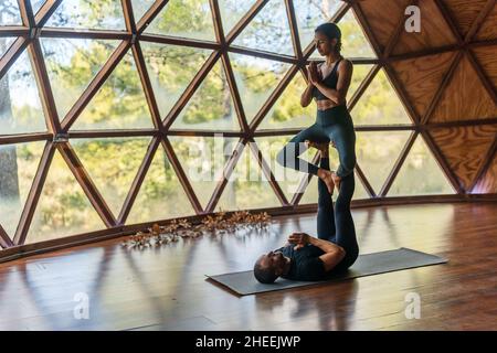 Full body of sporty couple in activewear performing yoga pose while him lying down with his legs stretched out and her doing namaste pose sitting on h Stock Photo