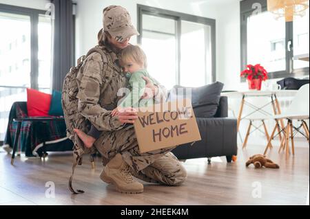 Side view of happy military female embracing son with Welcome Home tablet in light room after arriving home from mission Stock Photo