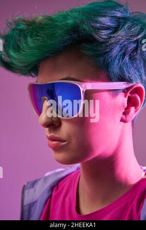 Close up of side view of stylish female with short green dyed hair in sunglasses with blue lens standing against purple in studio Stock Photo