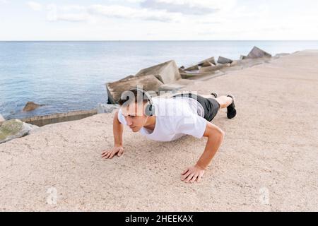 Strong sportive man listening to music in headphones while doing plank exercise on shore near rippling sea during intense workout Stock Photo