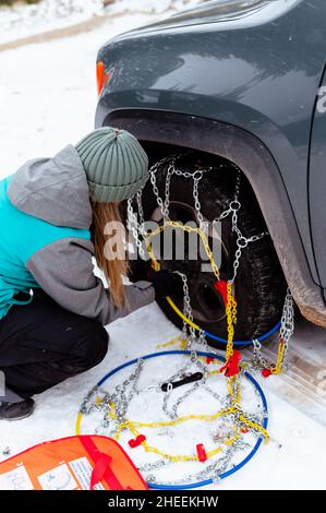 Side view of unrecognizable female wearing knitted hat and gloves and warm sportswear squatting and putting security snow chain on tire wheel of stuck Stock Photo