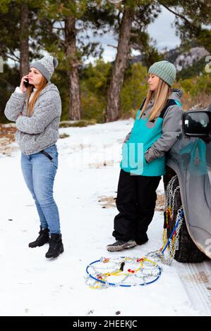 Full body of troubled Woman talking on smartphone with repairman of car service station while standing near female friend and stuck automobile in snow Stock Photo