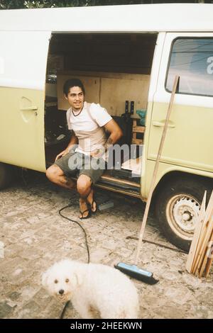 Full body of content Latin American male traveler looking away while sitting in camping van near dog during road trip Stock Photo