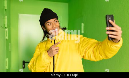 Asian male in outerwear and hat surfing internet on modern cellphone while standing near green wall and door on street Stock Photo