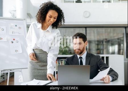 Two successful business partners of different nationalities working together on an important project, startup, in modern office, using laptop, brainstorming together, analyzing financial documents Stock Photo