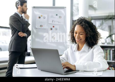 Positive young african american businesswoman, sales manager, sits in a modern office, working at laptop, smiles. On the background a male colleague ponders charts on a whiteboard, plans strategy Stock Photo