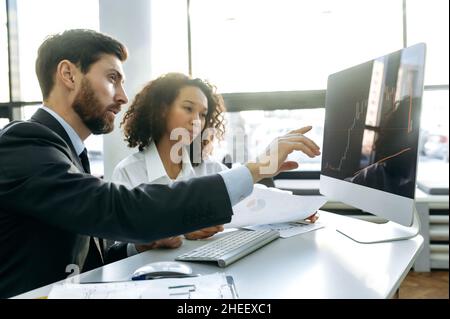 Successful Caucasian man and African American woman, crypto traders, sitting in an office, using a computer, looking at charts, analyzing the crypto market, planning a strategy for good profit Stock Photo