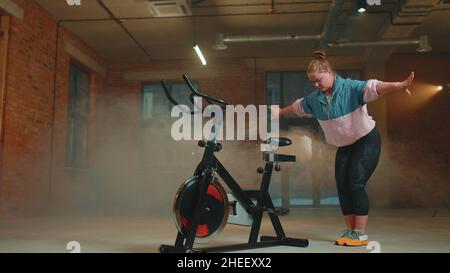 Athletic girl performing aerobic riding training exercises on cycling stationary bike in foggy gym Stock Photo