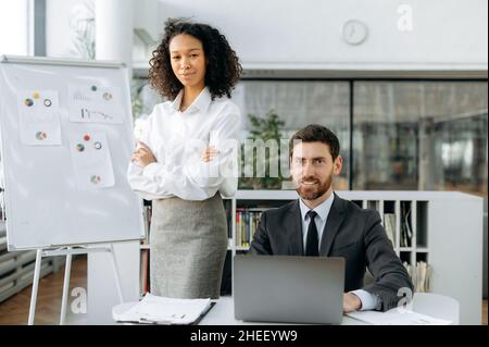Portrait of two business partners of different nationalities in a modern office. African american young woman and caucasian intelligent man dressed in formal clothes, looking at camera, smile friendly Stock Photo