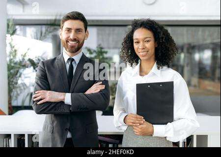 Friendly multiracial business colleagues, African American businesswoman, Caucasian businessman, company workers, standing in a modern office, looking at the camera, smiling Stock Photo