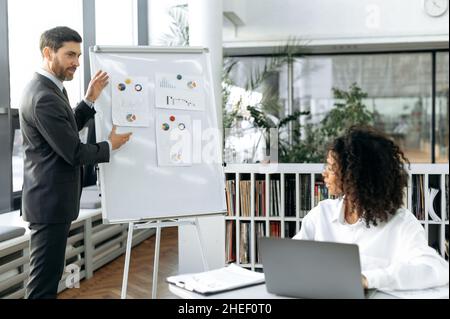 Working on a project, developing a financial strategy. Caucasian man, head of company, presenting strategy on whiteboard to his African American woman colleague, conducting brainstorm in modern office Stock Photo