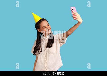 Smiling happy woman in party cone broadcasting livestream, recording video for blog during celebration her birthday, wearing white shirt. Indoor studio shot isolated on blue background. Stock Photo