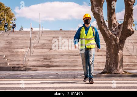 Latin worker with yellow hard hat, goggles, green vest and mouth guard, walking across the street on a pedestrian walkway. Stock Photo