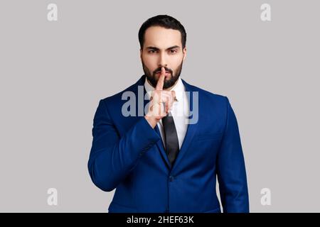 Shh, make silence pleas. Serious bearded businessman standing showing quiet gesture, saying hush, keep secret, wearing official style suit. Indoor studio shot isolated on gray background. Stock Photo