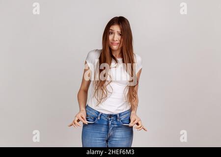 Portrait of upset poor woman with dark hair turning out empty pockets, worried about debts, no cash for living, wearing white T-shirt. Indoor studio shot isolated on gray background. Stock Photo