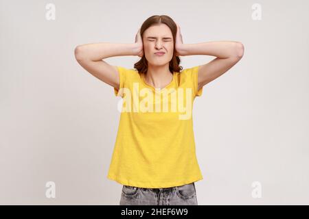 Don't want to listen. Beautiful irritated stressed out young woman in casual style clothing covering ears, keeping eyes closed, nervous breakdown. Indoor studio shot isolated on gray background. Stock Photo