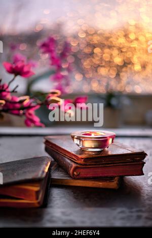 Wintertime with candle on stack of old books. Sunset window with orange glow, pink and fuchsia orchid and magnolia flowers. Aromatic candles, tea Stock Photo