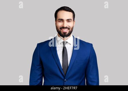 Cheerful bearded man with charming smile, standing and looking at camera, expressing positive emotions, wearing official style suit. Indoor studio shot isolated on gray background. Stock Photo