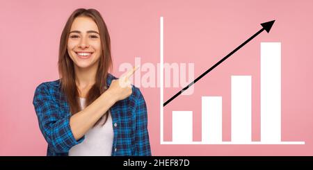 Portrait of happy joyful young woman standing, pointing aside and showing business growth graph. indoor studio shot isolated on pink background Stock Photo