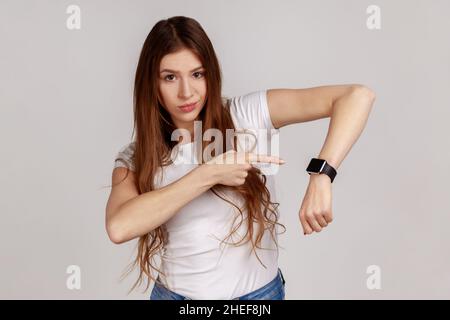 We have no time. Portrait of serious boss woman with dark hair pointing her finger at the smartwatch and getting angry, wearing white T-shirt. Indoor studio shot isolated on gray background. Stock Photo