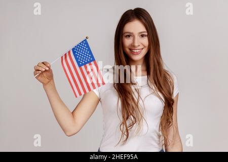 Portrait of smiling young adult woman holding USA national flag, celebrating national Independence Day - 4th july, wearing white T-shirt. Indoor studio shot isolated on gray background. Stock Photo