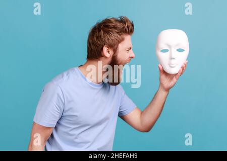 Portrait of bearded man screaming angrily, holding white mask in hands, hating his hidden personality, expressing negative, hatred. Indoor studio shot isolated on blue background. Stock Photo