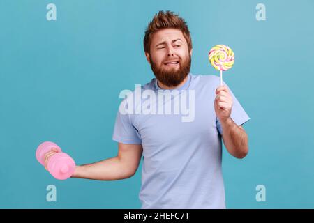 Portrait of funny handsome bearded man holding delicious lollypop and and raised up hand with pink heavy dumbbell, hard to do exercises. Indoor studio shot isolated on blue background. Stock Photo