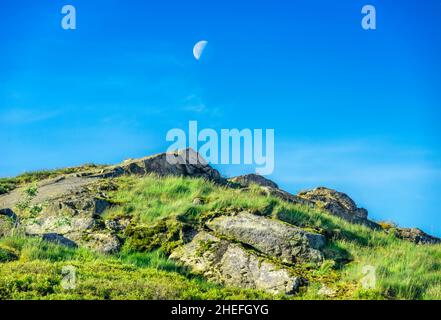 On a summer morning in June as the sun rises,the moon against blue sky,hanging over jagged rocks at Malvern Hills' highest peak,in early June. Stock Photo