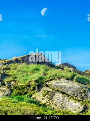On a summer morning in June as the sun rises,the moon against blue sky,hanging over jagged rocks at Malvern Hills' highest peak,in early June. Stock Photo