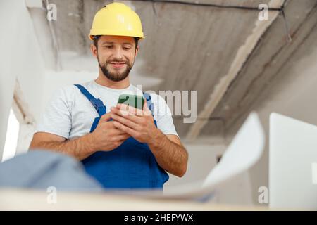 Focused repairman in workwear and hardhat smiling while using smartphone, standing indoors during renovation work Stock Photo