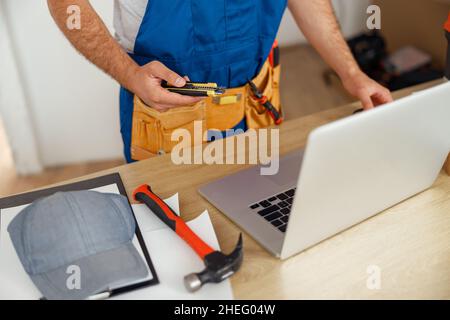 Cropped shot of repairman worker in uniform wearing tool belt, holding utitlity knife and using laptop while standing indoors Stock Photo