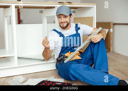 Smiling handyman, kitchen carpenter worker in overalls smiling at camera while installing kitchen furniture Stock Photo