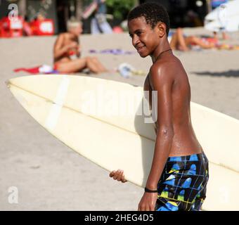 A dark skinned Indonesian boy walking on the beach and carrying a surfboard at Kuta Beach on the island of Bali in Indonesia. Stock Photo