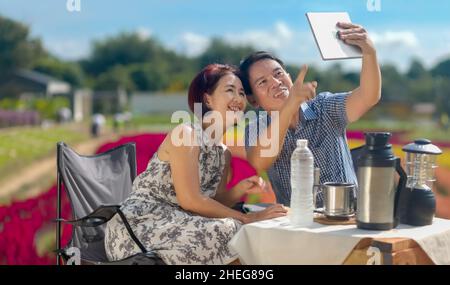 Happy middle aged couple selfie together while relaxed sitting in flower garden. Stock Photo