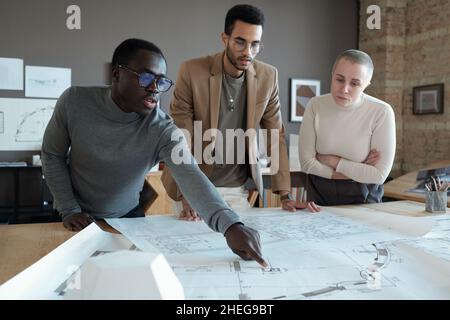 Young confident architect pointing at blueprint while bending over table and explaining sketch to colleagues at meeting Stock Photo