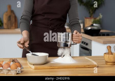 Young woman in apron sieving flour over wooden board while taking it out of white porcelain bowl with spoon Stock Photo