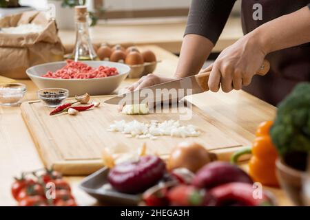 Hands of young female chopping fresh onion on wooden board while preparing italian pasta with vegetables and minced meat Stock Photo