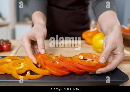 Hands of young female putting fresh chopped pepper on tray while standing by kitchen table and cooking vegetarian food Stock Photo