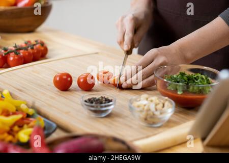 Hands of young female chopping fresh ripe cherry tomatoes for salad on wooden board while standing by kitchen table Stock Photo
